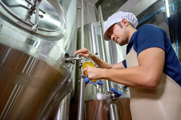 A young male brewer presses beer from a brewing keg into a glass. To determine the quality of beer and the taste and color of beer in the brewery industry.