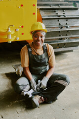 Vertical shot of happy young African American female technician wearing hardhat sitting on floor with wrench in hands smiling at camera