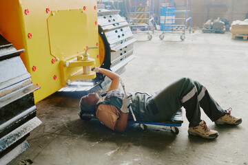 Young African American technician working in heavy machinery production factory, lying on creeper...