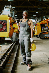 Vertical long of self-confident young African American female technician posing for camera with hardhat and wrench in hands at workplace in factory