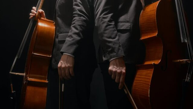 Cropped shot of two unrecognizable men wearing elegant black suits posing for camera on black background with cellos in hands