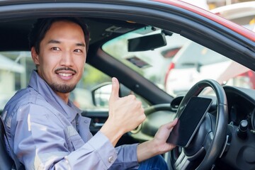 Asian male mechanic giving thumbs up while sitting in a car, smiling confidently. Represents customer satisfaction, professional service, and automotive repair success in a workshop setting.