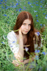 A girl with long curly red hair in white clothes sits in a meadow with cornflowers, summer day, cloudy warm weather