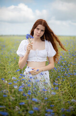 A girl with long curly red hair in white clothes stands in a meadow with cornflowers, holding a bouquet of cornflowers in her hands, summer day, cloudy warm weather
