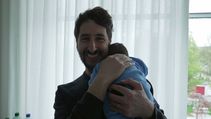 Father smiling while holding newborn baby in a blue onesie, standing by the window, capturing the joyful and tender moments of early bonding and the happiness of parenthood