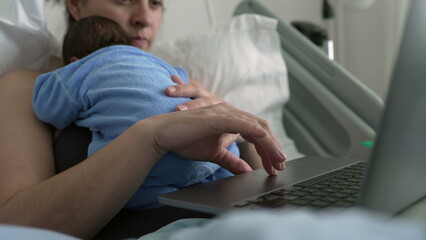 Mother balancing work and baby while working on a laptop, holding a newborn in a blue onesie. Close-up of multitasking hands, hospital bed, illustrating early motherhood and maternal care