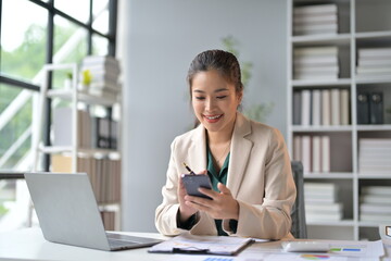 Businesswoman sitting at her desk using a smartphone and smiling. She is surrounded by paperwork and has a laptop in front of her
