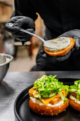 A person in black attire and gloves spreads cream cheese on a bagel with lettuce and tomato, preparing a sandwich.