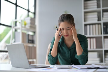 Obraz premium Asian accountant is suffering from a headache, touching her head with both hands, while sitting at her desk in the office