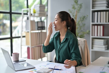 Obraz premium Young businesswoman is yawning while working at her desk in the office. She's surrounded by paperwork and appears to be tired