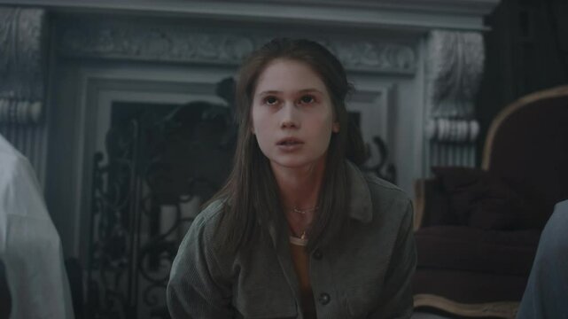 Chest up shot of teenagers holding friend who writhing in their hands while being possessed by demonic spirit, she suddenly opening black eyes and pronouncing prayer indoors