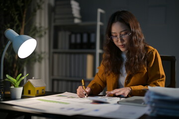 Young professional woman working overtime under a desk lamp, using a calculator and analyzing...