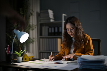 Determined young businesswoman works late crunching numbers for a project deadline in her small home office. Stress shows on her face as she focuses on her laptop, calculator, and paperwork