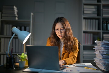 Tired businesswoman works late at night in her dimly lit office, focused on accounting tasks with a...