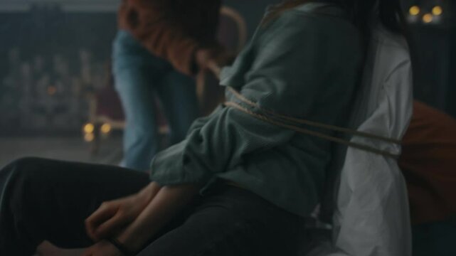 Side shot of pale demoniac teenage girl writhing on chair and pronouncing prayers while her panicking friends tying her up with rope after performing invoking ritual in old house