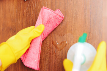 Woman in yellow gloves cleaning wooden surface with pink rag and green cleaning solution bottle
