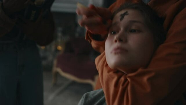 Closeup of unrecognizable guy drawing black cross with ember of palo santo wood stick on forehead of writhing demoniac girl while friend reading prayers from occult book, exorcism ceremony indoors