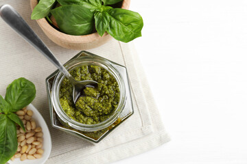 Tasty pesto sauce in jar, spoon, pine nuts and basil on white table, top view. Space for text