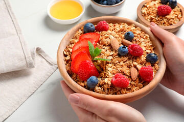 Woman holding bowl of tasty granola with berries, nuts and mint at white table, closeup