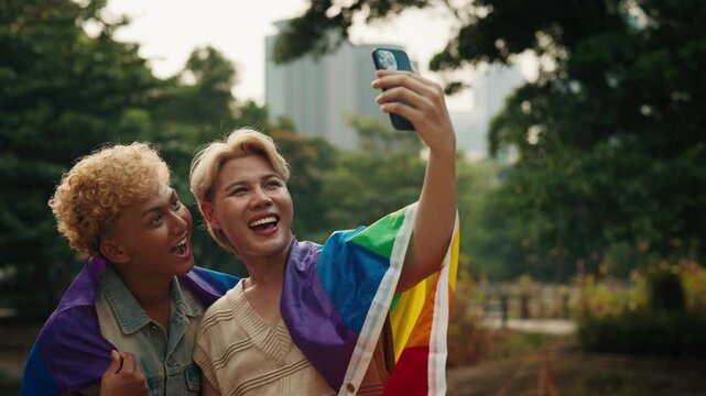 Happy young gays wrapped with rainbow flag make selfie in urban park. Handsome Asian men shows free love supporting LGBT community in pride month. Romantic homosexual relationship