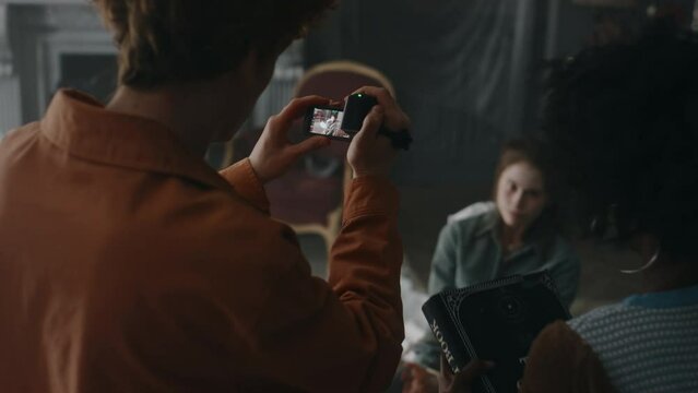 Back view of guy filming video on vhs camera of writhing demon possessed girl tied up to chair and of lady with occult book searching for prayers to perform exorcism ceremony in old derelict house