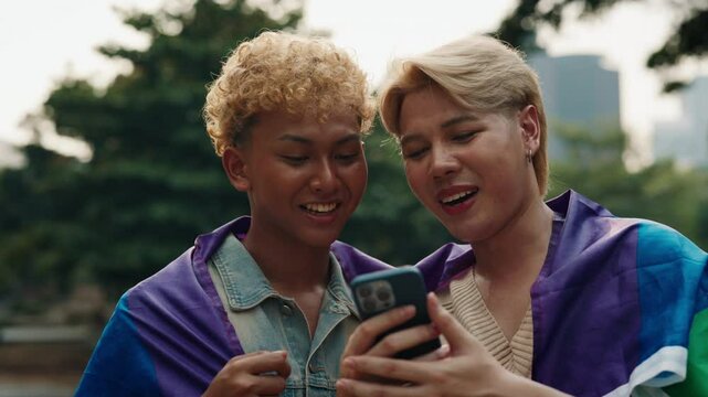 Positive homosexual couple with LGBT flag looks at photo with smartphone in urban park. Emotional Japanese men post on social media in pride month. Young gays visibility support