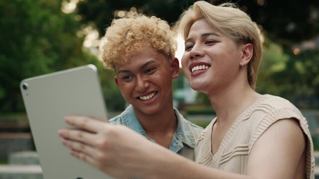 Handsome Asian gays talk to friends via call on tablet walking in green park slow motion. Joyful men romantic partners steam on social medial. LGBTQI pride month internet activity