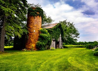 Old barn with a silo covered in ivy on a lush green farm under a partly cloudy sky. Michigan © Wirestock