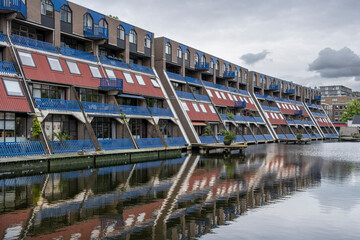 A row of modern, multi-story buildings with vibrant red and blue accents reflecting in a still canal, showcasing contemporary architecture alongside serene water imagery in Rotterdam