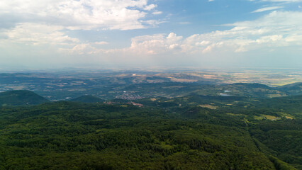 Drone flying above green mountain forest revealing vast landscape with small village under cloudy sky