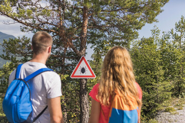 Couple with backpacks on a hiking route, standing in front of the tree and looking at a warning...