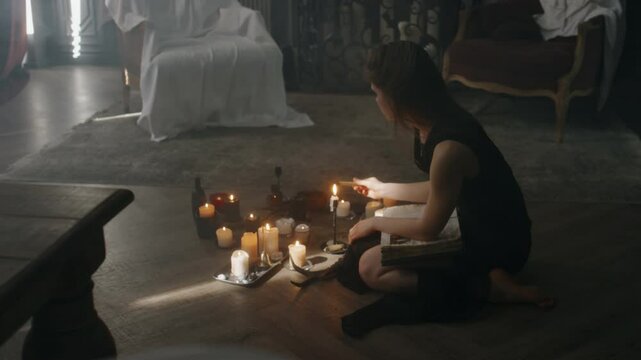 Side full shot of young Caucasian enchantress sitting on floor near multiple burning candles and lighting palo santo stick to clear old mansion from negative energy before performing dark ceremony