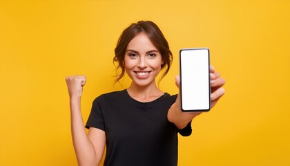 Naklejka premium Woman holds up a smartphone with a blank screen in one hand and raises her other fist in excitement, standing against a yellow background