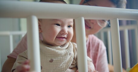 Obraz premium Adorable Baby infant Smiling Through playpen fence with grandmother in Background, Capturing showcasing a tender family Heartwarming Moment of Joy and Innocence in a Cozy Home Environment