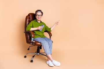 Photo portrait of lovely pensioner lady point empty space sit armchair dressed stylish green garment isolated on beige color background