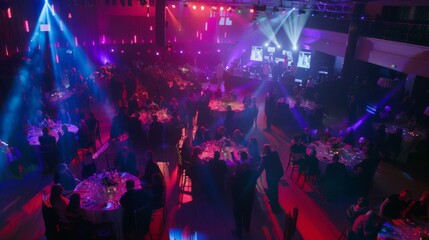A large reception hall with guests seated at tables under blue and pink lighting. The stage is set up with a band in the background.