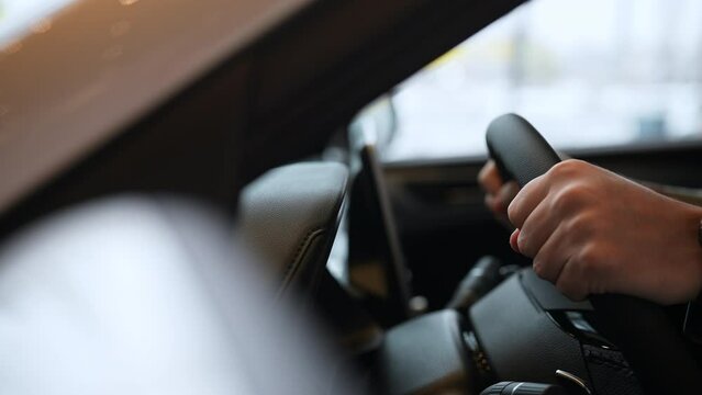 Close-up tracking shot of unrecognizable young man customer choosing new car in dealership center enjoying touching steering wheel. Male buyer sitting in a car interior at showroom, slow motion.