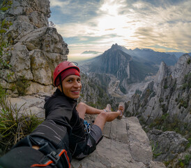 A climber wearing a red helmet takes a selfie on a rocky ledge with the stunning Monterrey mountain landscape in the background at sunrise. The morning sun casts a soft glow on the mountain