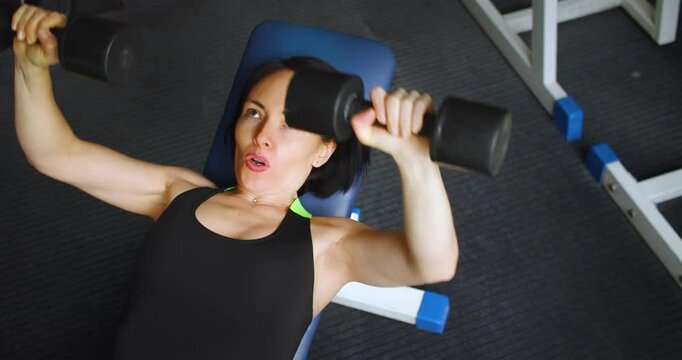 Top view of a middle aged fit Caucasian woman during gym workout. Bench press with dumbbells for chest. Physical education as a norm of life to maintain health. Slow motion 4k footage.