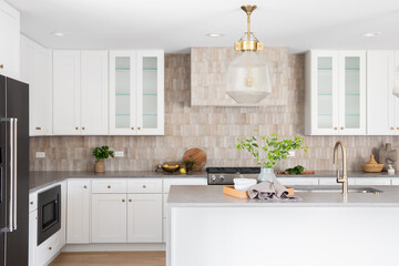 A kitchen detail with a grey stone countertop island, gold faucet, stainless steel appliances, brown tile backsplash, and white cabinets. No brands or labels.