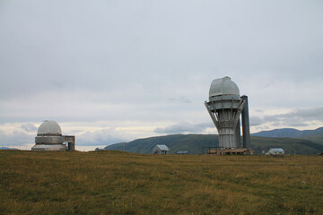 Two observatory telescopes stand on a grassy field against a backdrop of rolling hills and a cloudy...