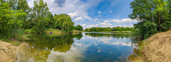 Panoramic view of the lake with deciduous trees on the banks. Non-urban landscape with picturesque nature countryside on a summer day
