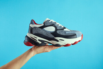 Hand holds hanging colored running sneakers by the laces on blue pastel background. Hand with a new casual shoe. Close up.