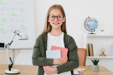 Confident smiling schoolgirl pupil student holding copybooks, standing at school, learning from...