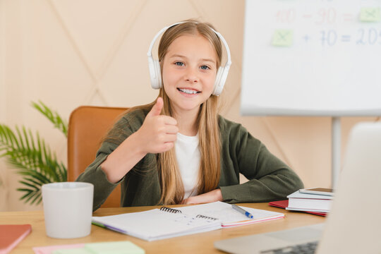 Smiling caucasian student pupil schoolgirl studying online, e-learning on laptop with headphones showing thumb up. Homeschooling concept. - Powered by Adobe