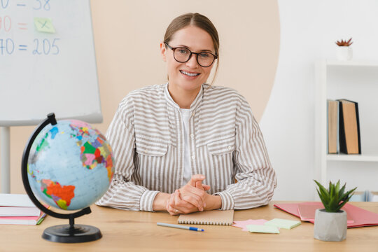 Young female maths geography teacher headmaster tutor student lector working at the desk looking at the camera in school classroom.