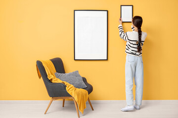 Young woman hanging picture frame on orange wall near armchair in stylish living room