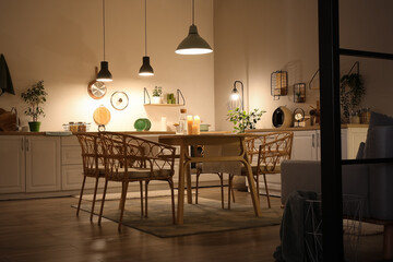 Interior of modern kitchen with glowing lamps, white counters, chairs and dining table at evening