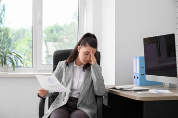 Young woman with paper sheet suffering from headache at workplace in office