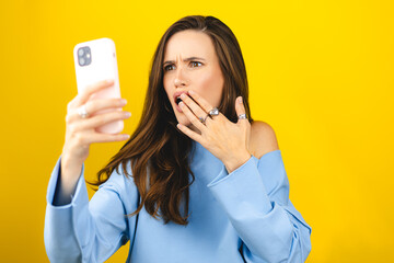 Mad and shock young brunette beautiful woman using mobile phone isolated on yellow background....